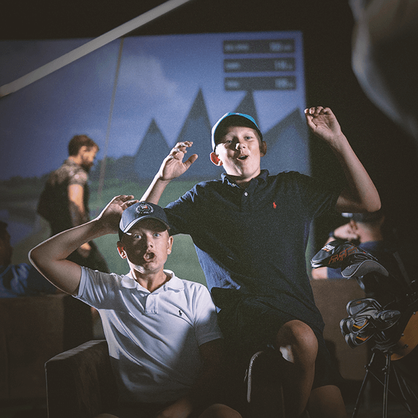 Two young golfers celebrating excitedly at an indoor golf simulator, highlighting the accessible fun at Sunday Nine Golf Club.