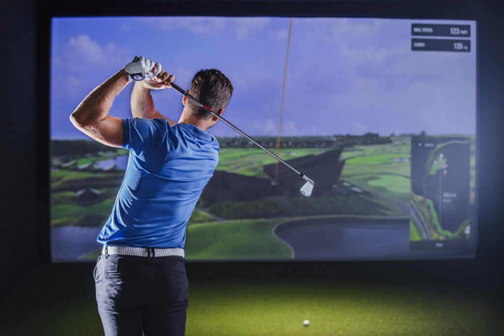 Golfer finishing a swing in a quiet simulator bay; dispersion chart on screen, notebook on table